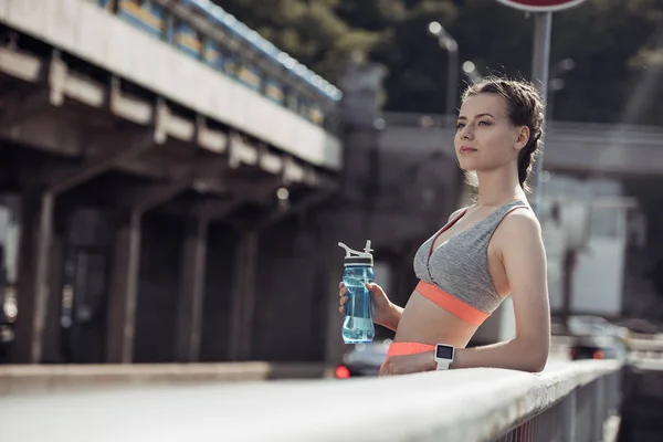 Приваблива спортивна дівчина тримає спортивну пляшку з водою і стоїть на перилах — стокове фото