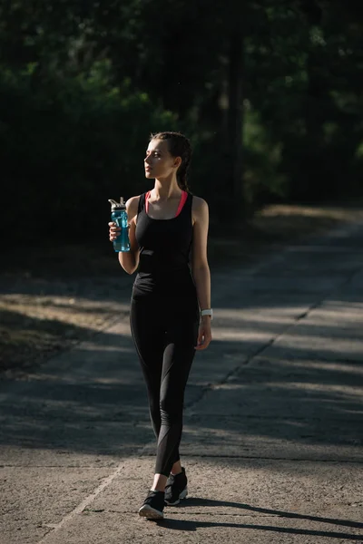 Donna atletica che tiene bottiglia sportiva e cammina sul sentiero nel parco — Foto stock