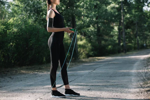 Vue recadrée d'une sportive avec un smartphone dans un brassard tenant une corde à sauter dans un parc — Photo de stock