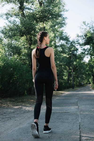 Vista posteriore della donna in abbigliamento sportivo ascolto musica con auricolari e smartphone nel parco — Foto stock