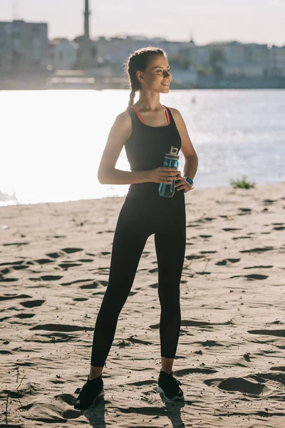 Hermosa deportista con agua en botella de deportes de pie en la playa - foto de stock