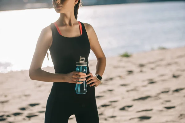 Vista recortada de chica atlética con botella de agua deportiva de pie en la playa - foto de stock