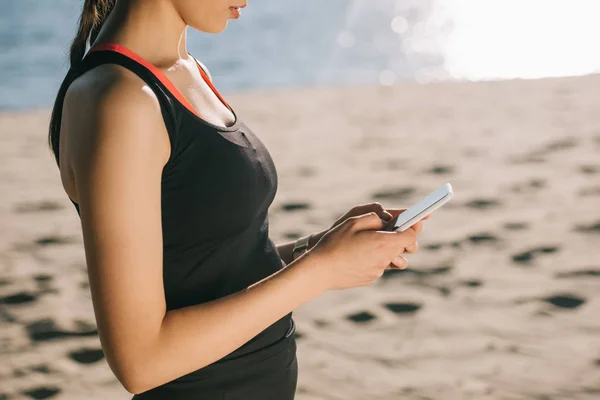 Cropped view of sportswoman listening music with earphones and using smartphone — Stock Photo