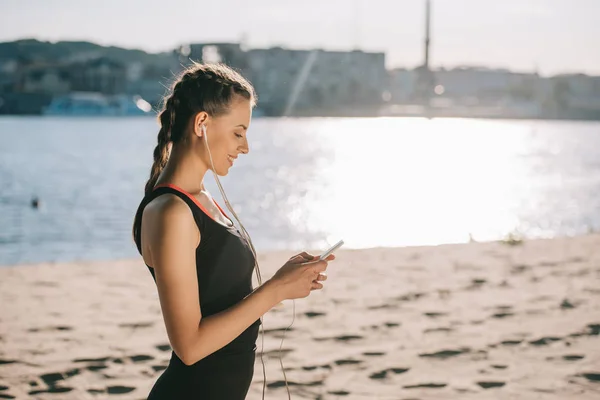 Smiling sporty girl listening music with earphones and using smartphone on beach with back light — Stock Photo