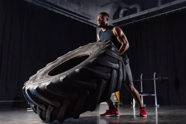 Vista de ángulo bajo del entrenamiento de hombre afroamericano muscular con neumático y mirando hacia otro lado - foto de stock