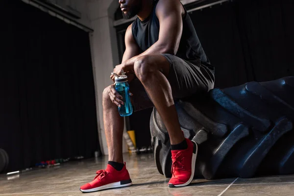 Recortado disparo de joven afroamericano deportista sentado neumático y la celebración de botella de agua en el gimnasio - foto de stock