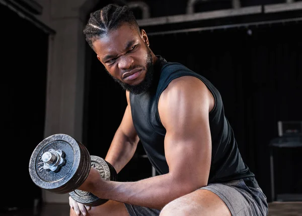 Jeune sportif afro-américain concentré s'entraînant avec haltère et regardant loin dans la salle de gym — Photo de stock