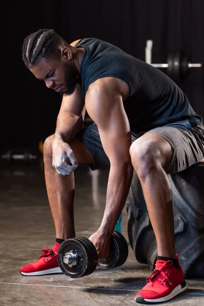 Athletic african american man sitting on tyre and exercising with dumbbell — Stock Photo