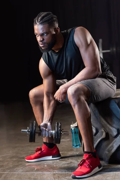 Sportif afro-américain musclé assis sur un pneu et faisant de l'exercice avec haltère — Photo de stock