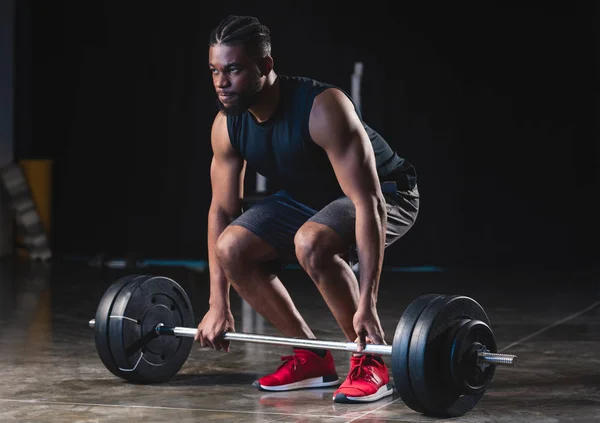 Enfocado muscular afroamericano deportista levantando la barra y mirando hacia otro lado en el gimnasio - foto de stock