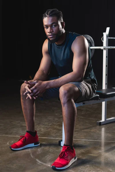 Visão completa do jovem esportista afro-americano sentado e olhando para a câmera no ginásio — Fotografia de Stock
