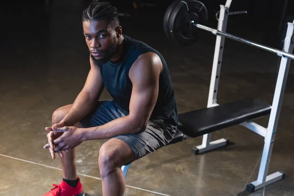 Vista de ángulo alto de joven afroamericano deportista sentado y mirando a la cámara en el gimnasio - foto de stock