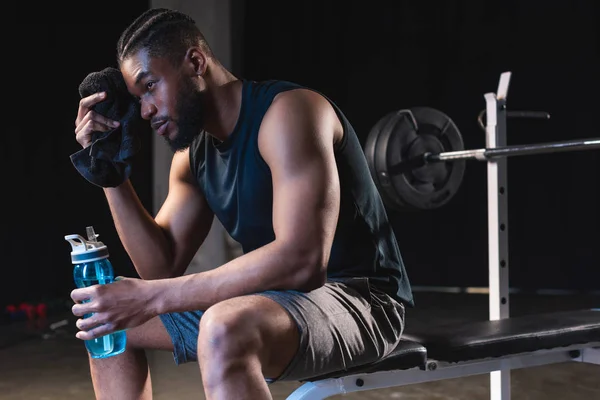 Tired african american sportsman holding towel and bottle of water while sitting in gym — Stock Photo