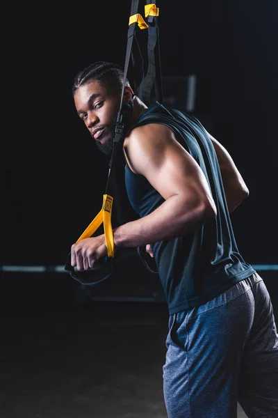 Joven deportista afroamericano mirando a la cámara mientras entrena con correas de fitness - foto de stock