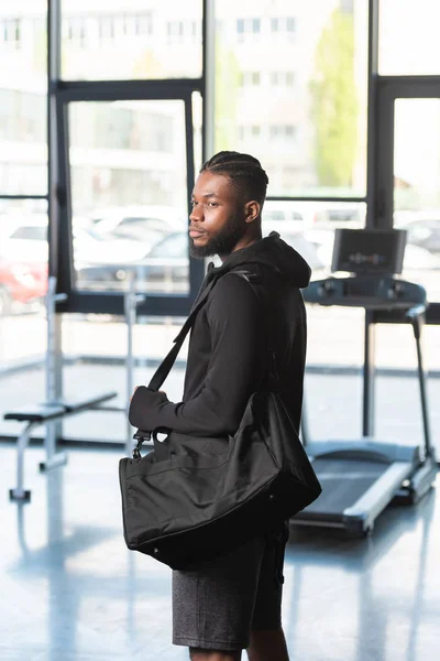 Guapo atlético afroamericano hombre en ropa deportiva bolsa de celebración y mirando hacia otro lado en el gimnasio - foto de stock