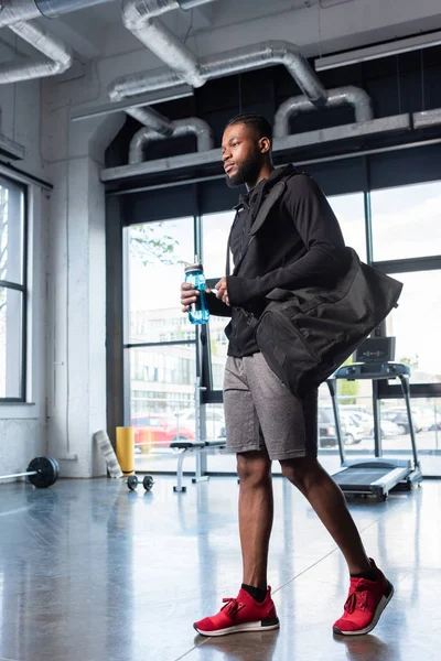 Low angle view of african american man with bag and bottle of water looking away in gym — Stock Photo