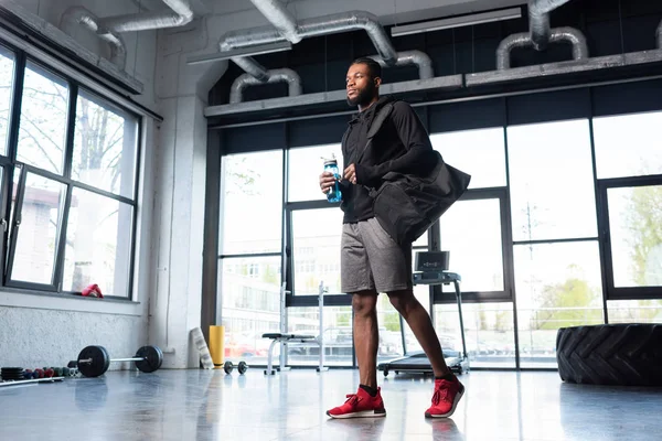 Vue à faible angle de l'homme afro-américain sportif tenant une bouteille d'eau et regardant loin dans la salle de gym — Photo de stock