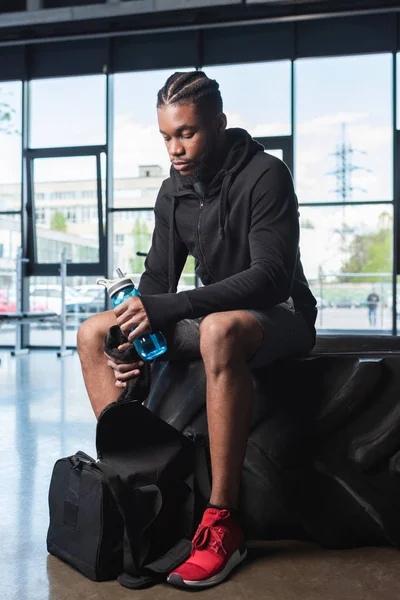 Joven deportista afroamericano sosteniendo botella de agua y toalla mientras está sentado en neumático en el gimnasio - foto de stock