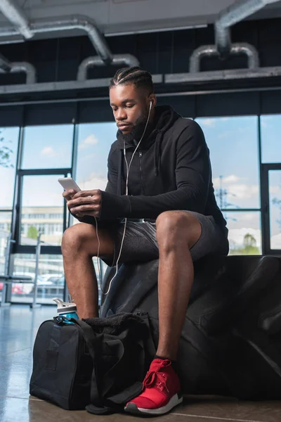 Young african american man in earphones sitting in tyre and using smartphone in gym — Stock Photo