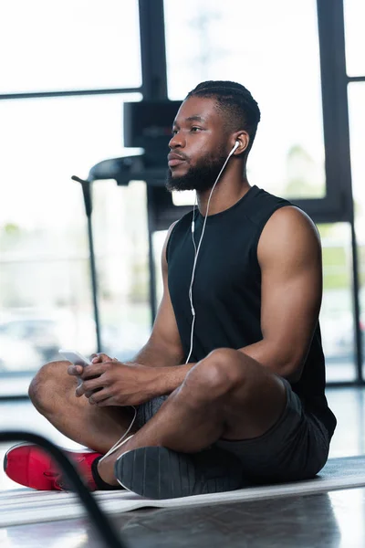 Deportista afroamericano pensativo en auriculares usando teléfono inteligente y mirando hacia otro lado mientras está sentado en la esterilla de yoga en el gimnasio - foto de stock