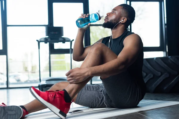 Jovem desportista afro-americano em fones de ouvido sentado no tapete de ioga e água potável no ginásio — Fotografia de Stock