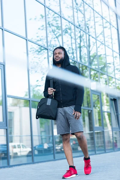 Vista de ángulo bajo del joven afroamericano en ropa deportiva caminando con bolsa en la calle - foto de stock