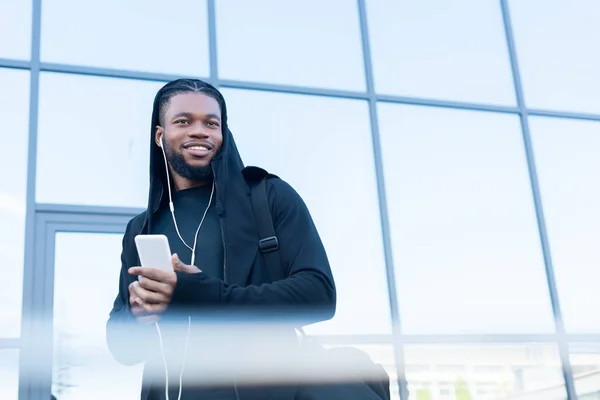 Enfoque selectivo de guapo sonriente hombre afroamericano en auriculares usando teléfono inteligente en la calle - foto de stock