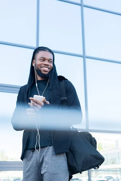 Foco seletivo de sorrir homem americano africano usando smartphone e olhando para longe — Fotografia de Stock