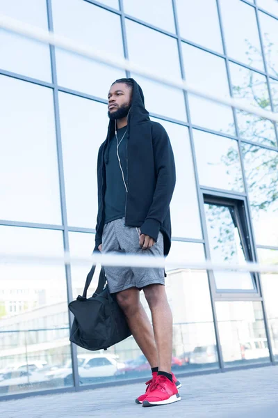 Vista basso angolo di giovane uomo afroamericano in auricolari in possesso di borsa sportiva e guardando altrove — Foto stock