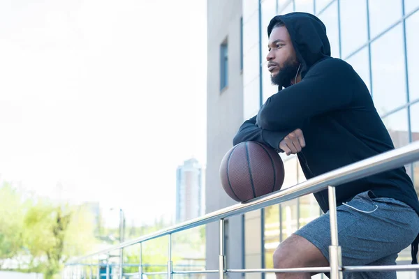Vista lateral del hombre afroamericano pensativo apoyado en barandillas con pelota de baloncesto y mirando hacia otro lado - foto de stock