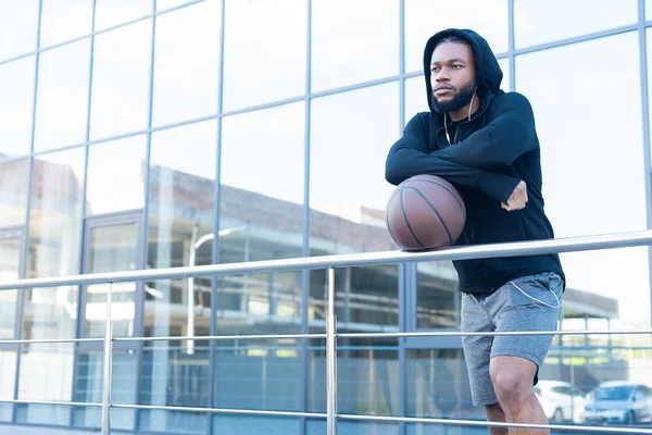 Homem americano africano pensativo em fones de ouvido inclinando-se para grades com bola de basquete e olhando para longe — Fotografia de Stock