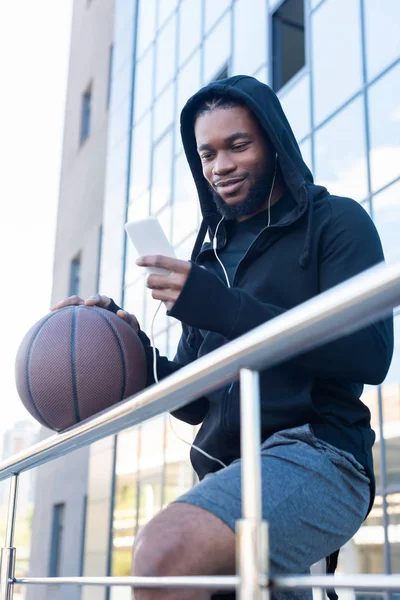 Sonriente hombre afroamericano en auriculares usando teléfono inteligente mientras sostiene la pelota de baloncesto en la calle - foto de stock