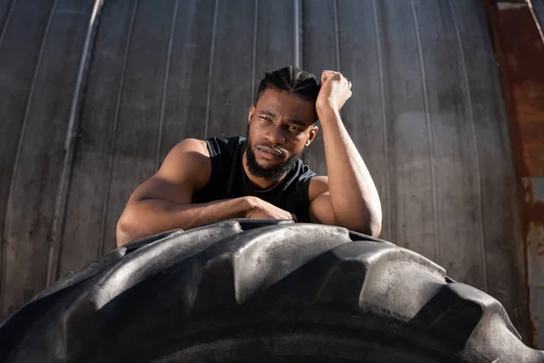 Beau jeune sportif afro-américain penché sur le pneu et regardant la caméra — Photo de stock