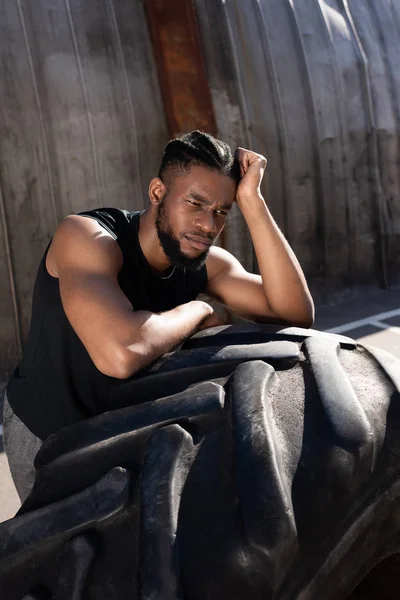 Pensive african american sportsman leaning at tyre and looking away on street — Stock Photo