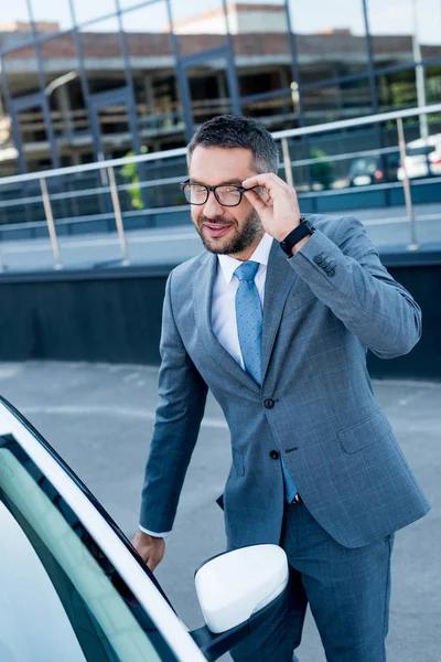 Улыбающийся бизнесмен в очках открывает дверь машины на улице — стоковое фото