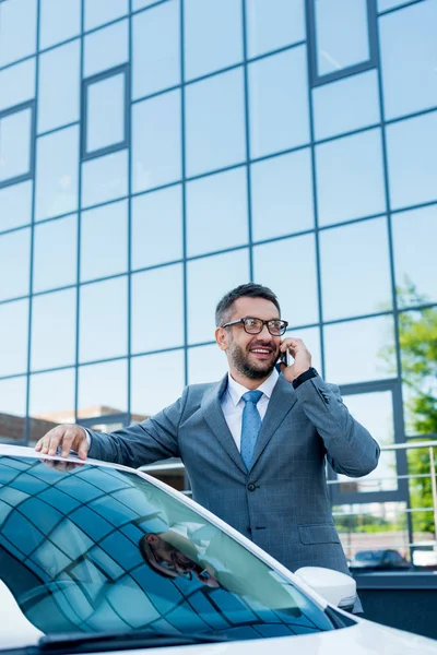 Porträt eines lächelnden Geschäftsmannes, der auf der Straße am Auto steht und mit dem Smartphone spricht — Stockfoto