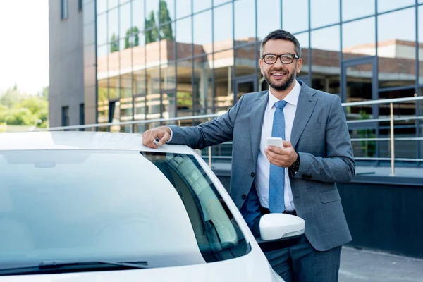 Портрет усміхненого бізнесмена зі смартфоном, що стоїть на машині на вулиці — стокове фото