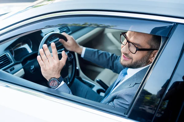 Homme d'affaires souriant en costume et lunettes saluant quelqu'un tout en conduisant une voiture — Photo de stock