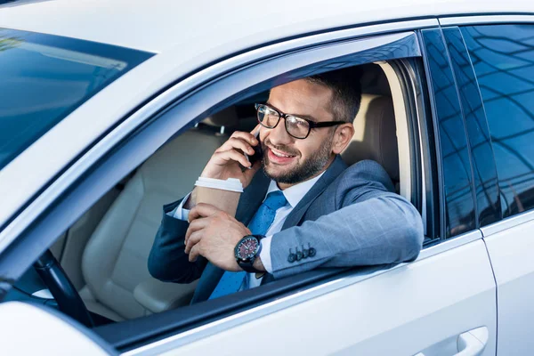 Homem de negócios sorridente com café para ir falar no smartphone no carro — Fotografia de Stock
