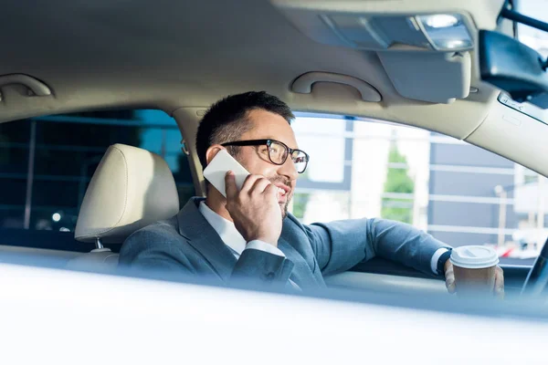 Vista lateral de hombre de negocios sonriente con café para ir a hablar en el teléfono inteligente en el coche - foto de stock