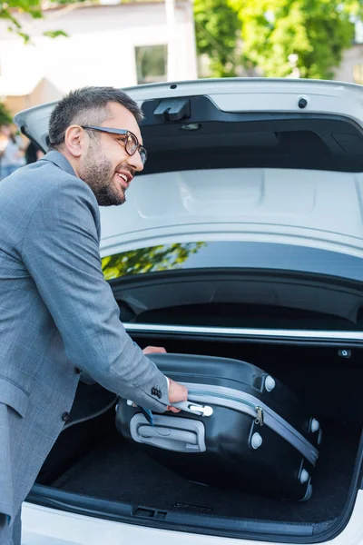 Seitenansicht eines lächelnden Geschäftsmannes, der sein Gepäck auf dem Parkplatz ins Auto legt — Stockfoto