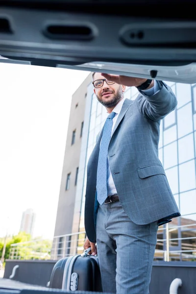 Businessman in eyeglasses with luggage standing at car on parking — Stock Photo