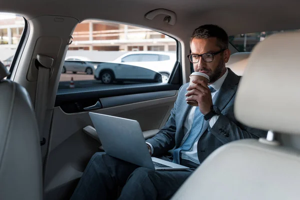 Businessman in eyeglasses with coffee to go using laptop in car — Stock Photo