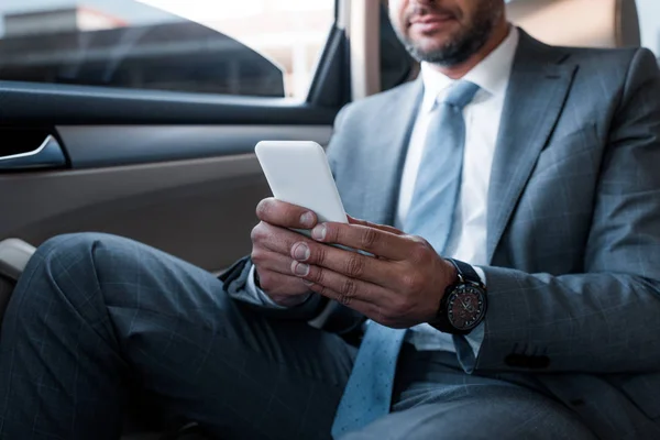 Vista parcial del hombre de negocios usando el teléfono inteligente mientras está sentado en el asiento trasero en el coche - foto de stock