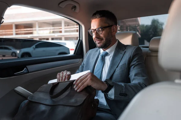 Hombre de negocios en traje poniendo papeles en la bolsa mientras está sentado en el asiento trasero en el coche - foto de stock