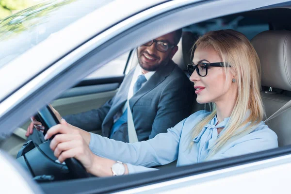Vista lateral de mujer de negocios rubia en gafas de conducir coche con colega cerca de - foto de stock