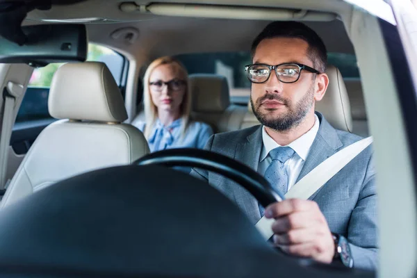 Selective focus of businessman in eyeglasses driving car with colleague sitting behind on passengers seat — Stock Photo
