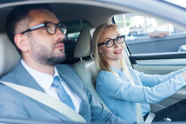 Side view of blonde businesswoman in eyeglasses driving car with colleague near by — Stock Photo