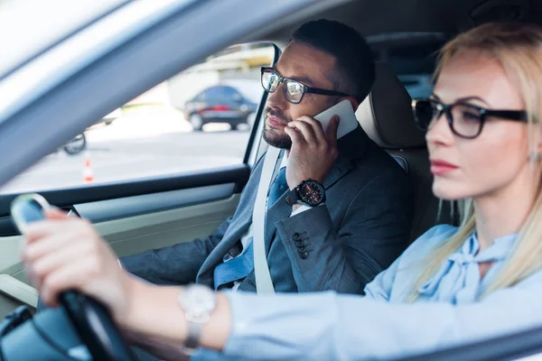 Foco seletivo de empresária dirigindo carro enquanto colega falando no smartphone perto de no assento dos passageiros — Fotografia de Stock