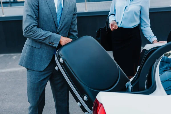 Recortado disparo de hombre de negocios poner equipaje en el coche con colega cerca de - foto de stock